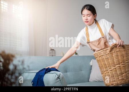 Beautiful young housekeeper maid doing housework holds basket wood of clean messy dirty clothes at home, Housewife woman holding basket with heap of d Stock Photo