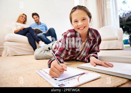 Homework done equals relaxing time. a little girl lying on the living room floor doing homework with her parents in the background. Stock Photo