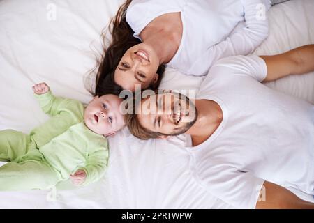 He always wants to be close to mommy and daddy. a young couple lying on the bed with their baby boy. Stock Photo
