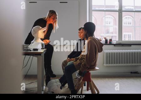 Smiling teacher discussing with multiracial students sitting in front of illuminated social robot in innovation lab Stock Photo