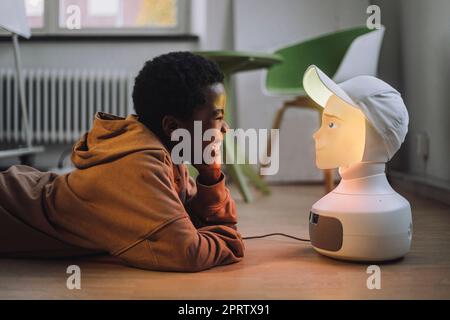 Cheerful boy in front of illuminated AI robot while lying on floor in innovation lab Stock Photo