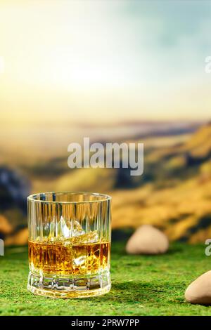 Glass of smoky whiskey placed on green moss with defocused mountain landscape on background Stock Photo