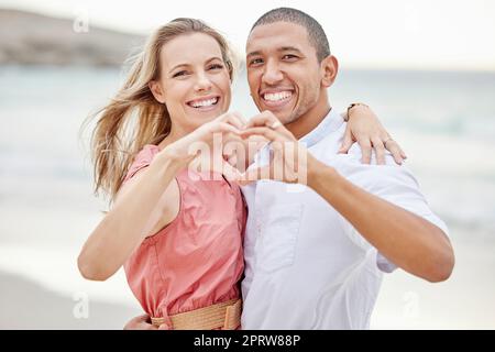 Happy interracial couple show heart sign with their hands and enjoying fresh air on vacation at the beach while bonding. Portrait of couple hugging at ocean with smile and love together on holiday Stock Photo