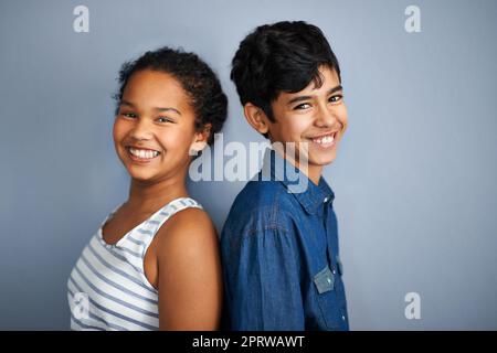 Back to back besties. A cute brother and sister standing isolated on grey. Stock Photo