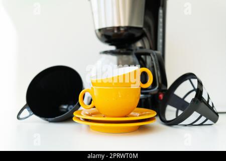 An automatic drip coffee maker stands with a yellow cup on a white table. Black modern drip coffee pot. Electric kitchen small household appliances. Appliances. Stock Photo