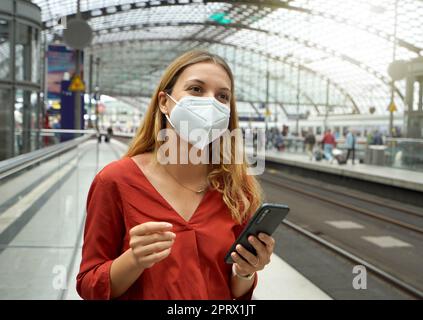 Traveler woman wearing KN95 FFP2 face mask at the airport train station. Young caucasian woman waiting shuttle train to the airport. Stock Photo