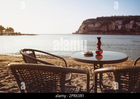 empty cafe table and chairs on sandy beach with ashtray and candle light in front of sea golf with clear sky before sunset Stock Photo