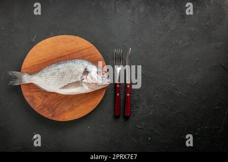 top view raw sea fish on round wood board fork and knife on black background copy place Stock Photo