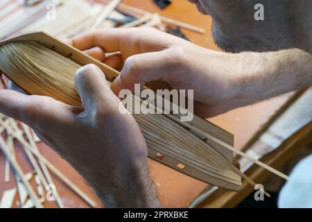 Hands of man gluing plywood details for ship model with glue, holding with fingers. Process of building toy ship, hobby, handicraft. Table with variou Stock Photo