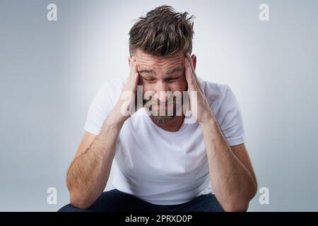 Does anyone have a headache tablet. a young man with a headache holding his head while sitting on a chair in the studio Stock Photo