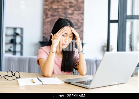 Tired woman sitting at the laptop after hard day, overworked female entrepreneur, wrinkled forehead, holding hand with the head, has a headache, a lot of information, glasses and notebook on the desk Stock Photo