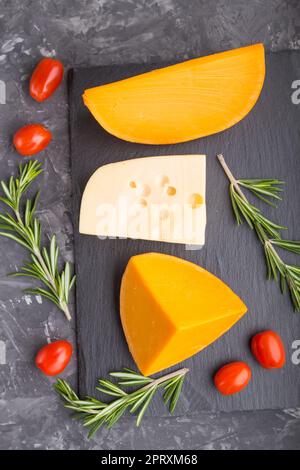 Various types of cheese with rosemary and tomatoes on black slate board on a black concrete background. Top view, flat lay, close up. Stock Photo