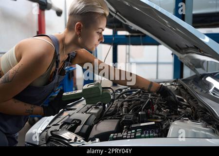 Young woman works under the hood of a car with a special scanner, female in work overalls Stock Photo