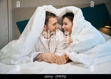 Young spouses have fun chatting under covers on large bed Stock Photo