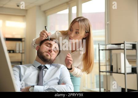 Young woman drawing on colleague's face while he sleeping in office. Funny joke Stock Photo