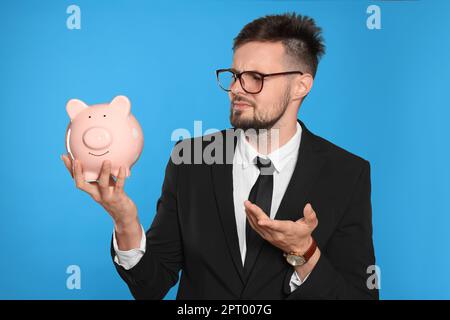 Confused businessman with piggy bank on light blue background Stock Photo