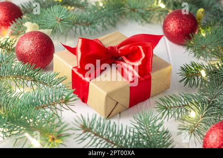 Gift wrapped in craft paper and red ribbon with green branches of Christmas tree with LED light bulbs, garland and shiny balls on a white wooden backg Stock Photo