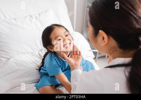 little asian girl showing teeth to blurred doctor with tongue depressor in hospital ward,stock image Stock Photo