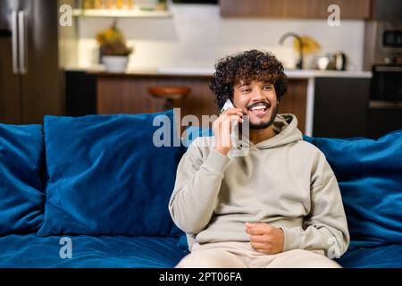 Portrait of carefree young Indian guy sitting on sofa and talking on smartphone, serene curly ethnic man in casual hoodie in relaxed atmosphere spending leisure at home, enjoying phone conversation Stock Photo