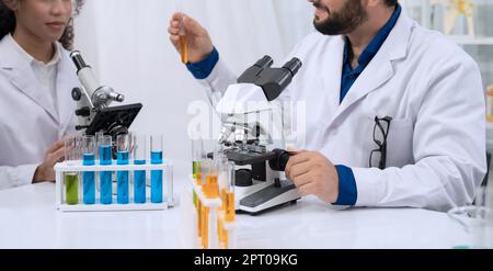 Two scientist researchers wearing white coat and working in laboratory. Man holding test tube with yellow liquid and using microscope. Chemistry, medi Stock Photo