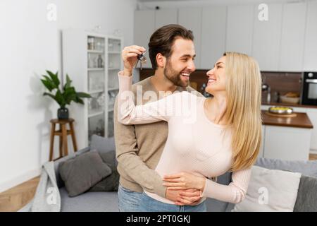 Happy young caucasian married couple holding key ring in hand, hugging and looking at each other with love, while standing in the modern kitchen of their new home. Excited owners of a family apartment Stock Photo