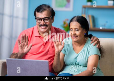 Happy Indian elderly couple talking on video calling using laptop while on sofa at home - concept of online communication, retirement lifestyle and Stock Photo