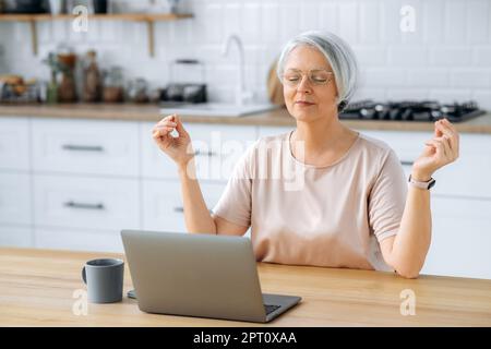 Calm relaxed satisfied middle aged caucasian grey haired woman with glasses, sitting at home in the kitchen at the table with a laptop, relaxing while working online, meditating with her eyes closed Stock Photo