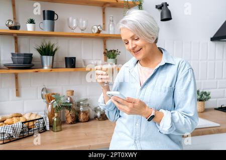 Satisfied caucasian middle aged gray haired woman, stand at home in the kitchen, holds her smartphone and a glass of coffee, chatting in social networks, reading news, makes an online order, smiles Stock Photo