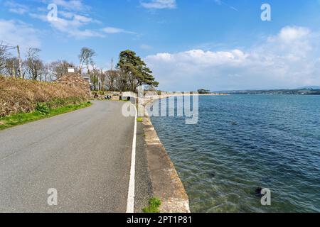 A tranquil and scenic view of the Menai Straits near Anglesey, Wales with blue sky, white clouds, green land and trees beside a winding coastal road. Stock Photo