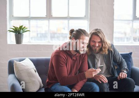 twin brothers using smartphone browsing internet on mobile phone sitting on sofa at home. Stock Photo