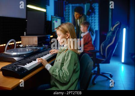 School children creating music at sound record studio, focus on girl playing synthesizer Stock Photo