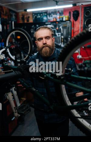 Vertical portrait of brutal cycling repairman standing by bicycle in repair workshop with dark interior, looking at camera. Concept of repair and Stock Photo