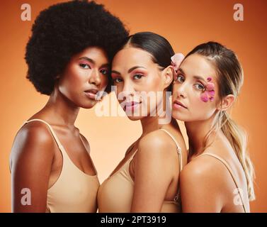 Women, flowers and beauty in makeup, diversity and models on orange studio background. Cosmetic, skincare and happy, young and elegant female friends Stock Photo