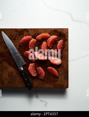 Peeled and sliced, divided in sections grapefruit on a wooden board with a knife next to it. The board rests on the marble countertop Stock Photo