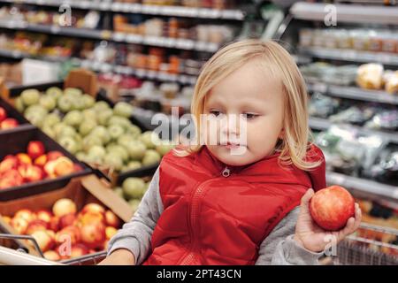 Adorable toddler girl sitting in shopping cart in food fruit store or supermarket. Portrait little cute kid going shopping and buying fresh fruits and Stock Photo