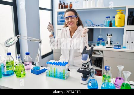 Young hispanic woman working at scientist laboratory screaming proud, celebrating victory and success very excited with raised arm Stock Photo