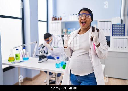Young hispanic woman expecting a baby working at scientist laboratory amazed and surprised looking up and pointing with fingers and raised arms. Stock Photo