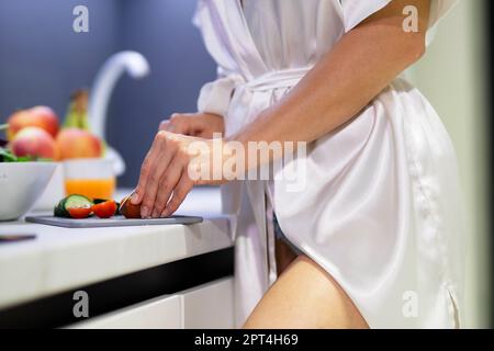 Close up shot of young female in white robe preparing breakfast in kitchen. Caucasian woman slicing fresh cucumbers and cherry tomatoes on cutting Stock Photo