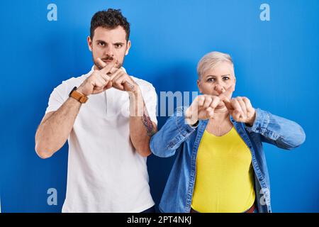 Young brazilian mother and son standing over blue background rejection expression crossing fingers doing negative sign Stock Photo
