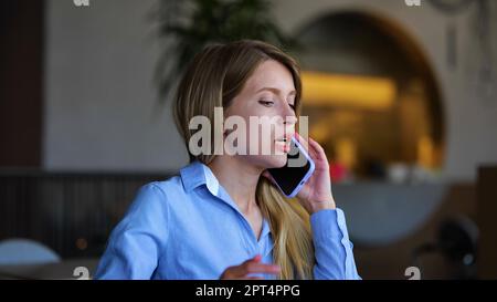 Camera moving back from young pretty female sitting in restaurant and looking at laptop screen while calling on cellphone. Beautiful business lady Stock Photo