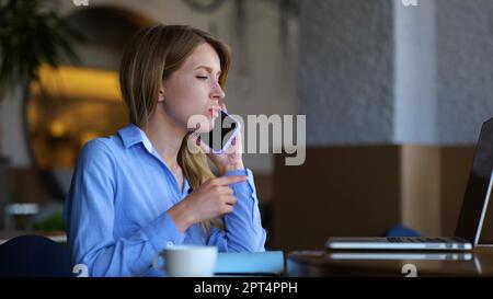Beautiful young female speaking on smartphone and using laptop while sitting in restaurant. Close up of happy pretty businesswoman chatting on mobile Stock Photo