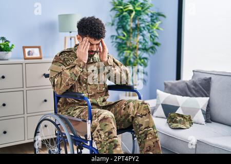 Arab man wearing camouflage army uniform sitting on wheelchair with hand on head for pain in head because stress. suffering migraine. Stock Photo