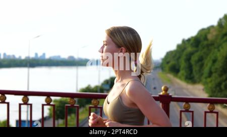Close up of Caucasian pretty sportswoman in sportswear running in street on the bridge. Side view. Young female athlete jogging outside with view on a Stock Photo