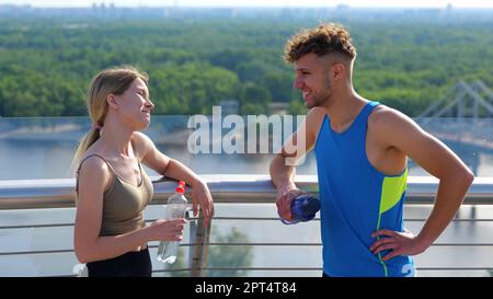 Close up portrait of Caucasian young sportswoman speaking with handsome sportsman outside in city with bottle of water. Male and female athletes Stock Photo