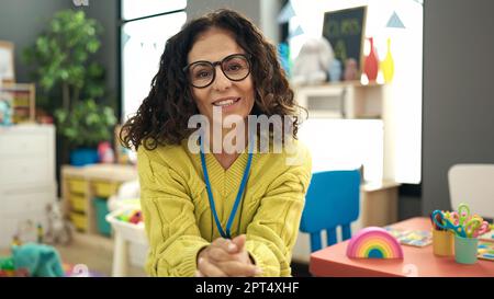 Middle age hispanic woman preschool teacher smiling confident sitting on chair at kindergarten Stock Photo