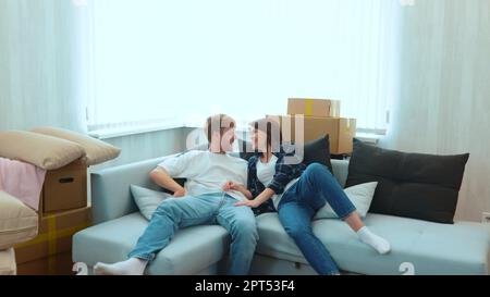Camera approaching joyful male and pretty female smiling jumping on a sofa in light room with many cardboard boxes. Close up of happy boyfriend and Stock Photo