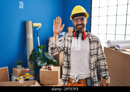 Young hispanic man with beard working at home renovation waiving saying hello happy and smiling, friendly welcome gesture Stock Photo