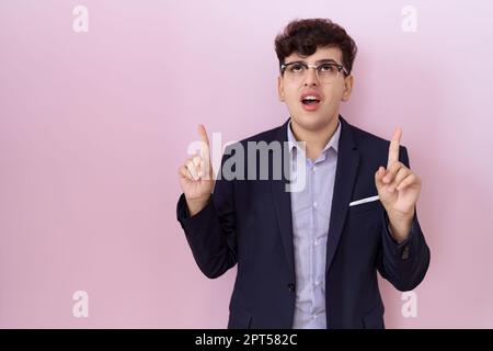 Young non binary man with beard wearing suit and tie amazed and surprised looking up and pointing with fingers and raised arms. Stock Photo