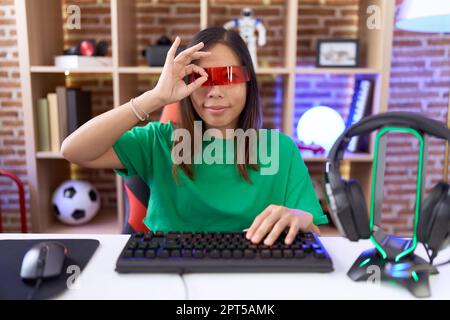 Middle age chinese woman wearing virtual reality glasses doing ok gesture with hand smiling, eye looking through fingers with happy face. Stock Photo
