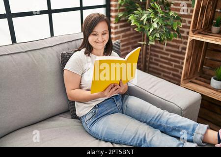 Down syndrome woman reading book sitting on sofa at home Stock Photo